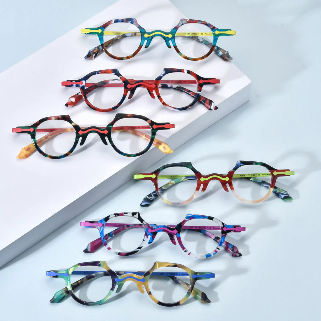 Group image of VoyueLens™ VL19341 designer round eyeglasses featuring all six color variants: Amber Crystal, Cobalt Blue, Orchid Purple, Champagne Clear, Tortoise Green Flame, and Ink Black Garnet. Each lightweight acetate frame showcases bold artistic patterns with glossy finishes, presented on a pastel background for a modern and expressive style overview.