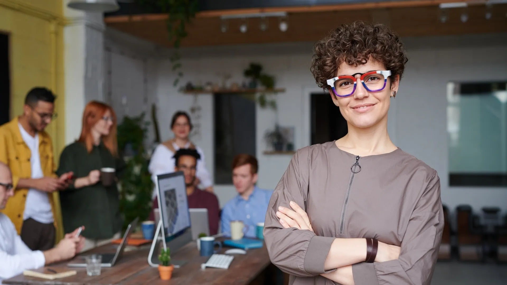 Confident woman wearing bold VoyueLens™ glasses in a modern office setting, with team collaborating in the background
