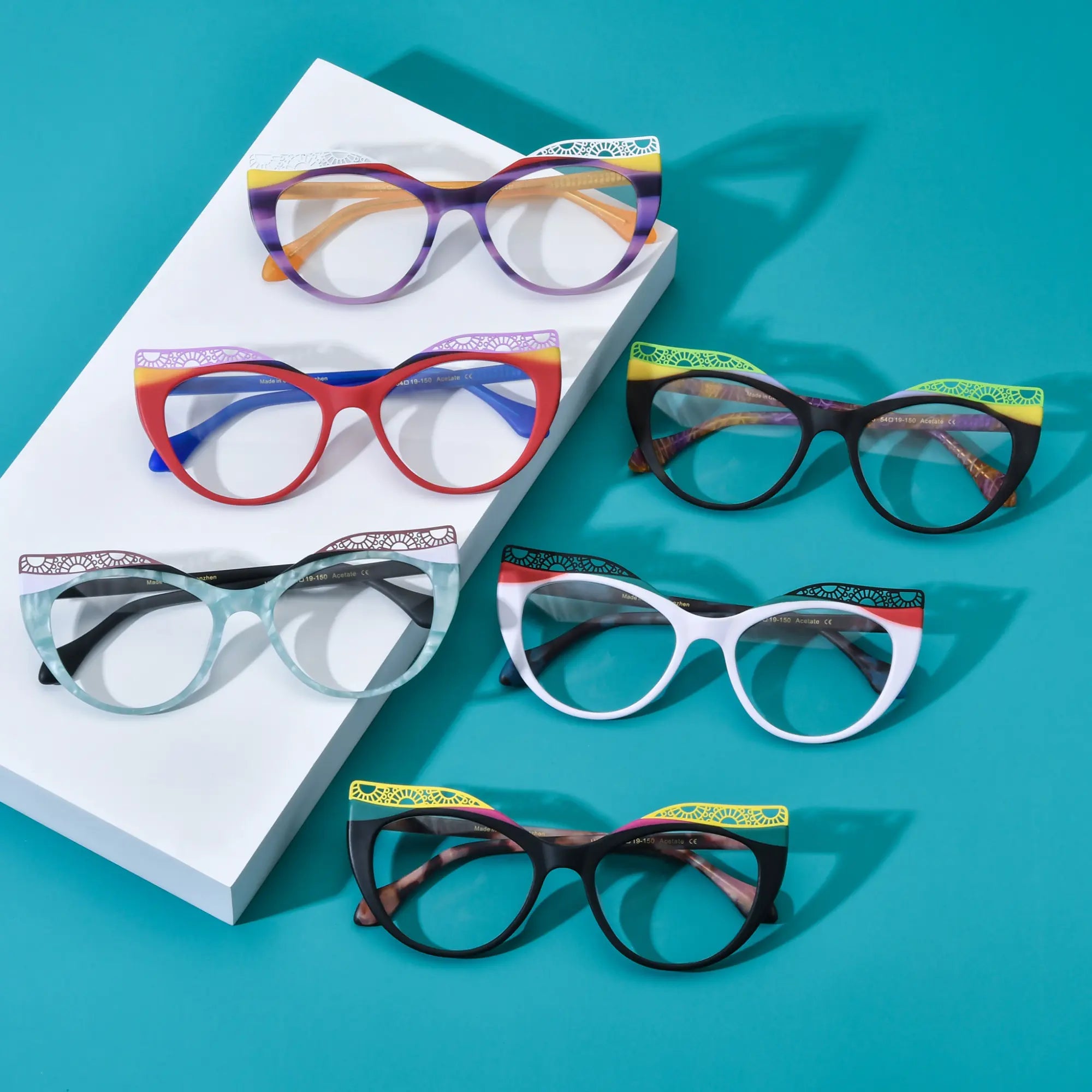 Group display of VoyueLens™ VL19365 cat-eye eyeglasses in six vibrant gradient variants: Amethyst Sunrise, Scarlet Horizon, Ice Coral, Aqua Frost, Citrus Tortoiseshell, and Crimson Nebula. Bold butterfly frames with lace-patterned upper edges shown on a turquoise background.
