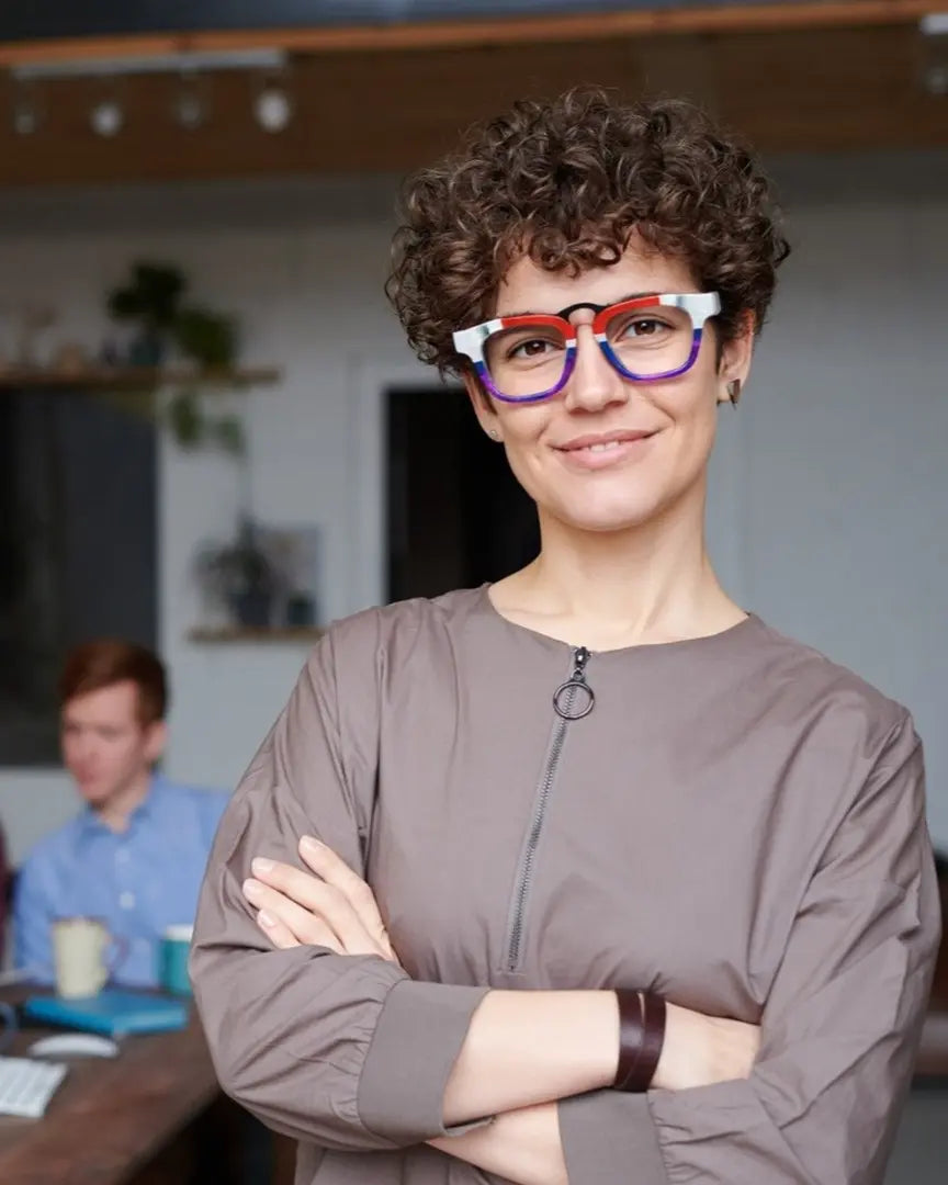 Confident woman wearing bold VoyueLens™ glasses in a modern office setting, with team collaborating in the background