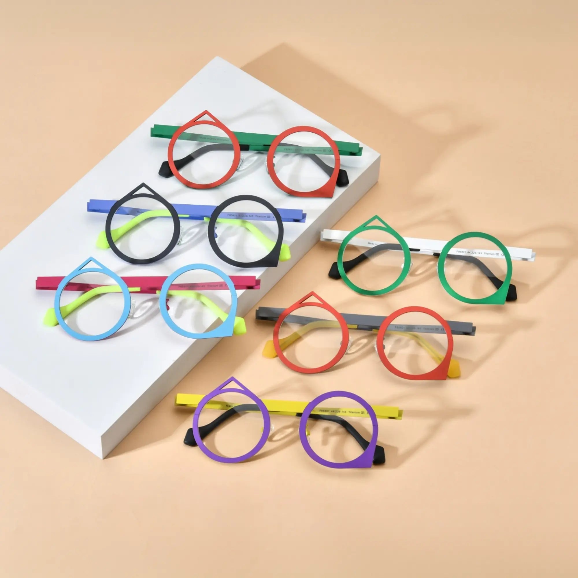 Top-down overview of all six color variants of VoyueLens™ VL185801 geometric titanium eyeglasses displayed on a beige background, showcasing red-green, purple-yellow, red-grey, green-white, blue-pink, and black-blue artistic polygonal frames.