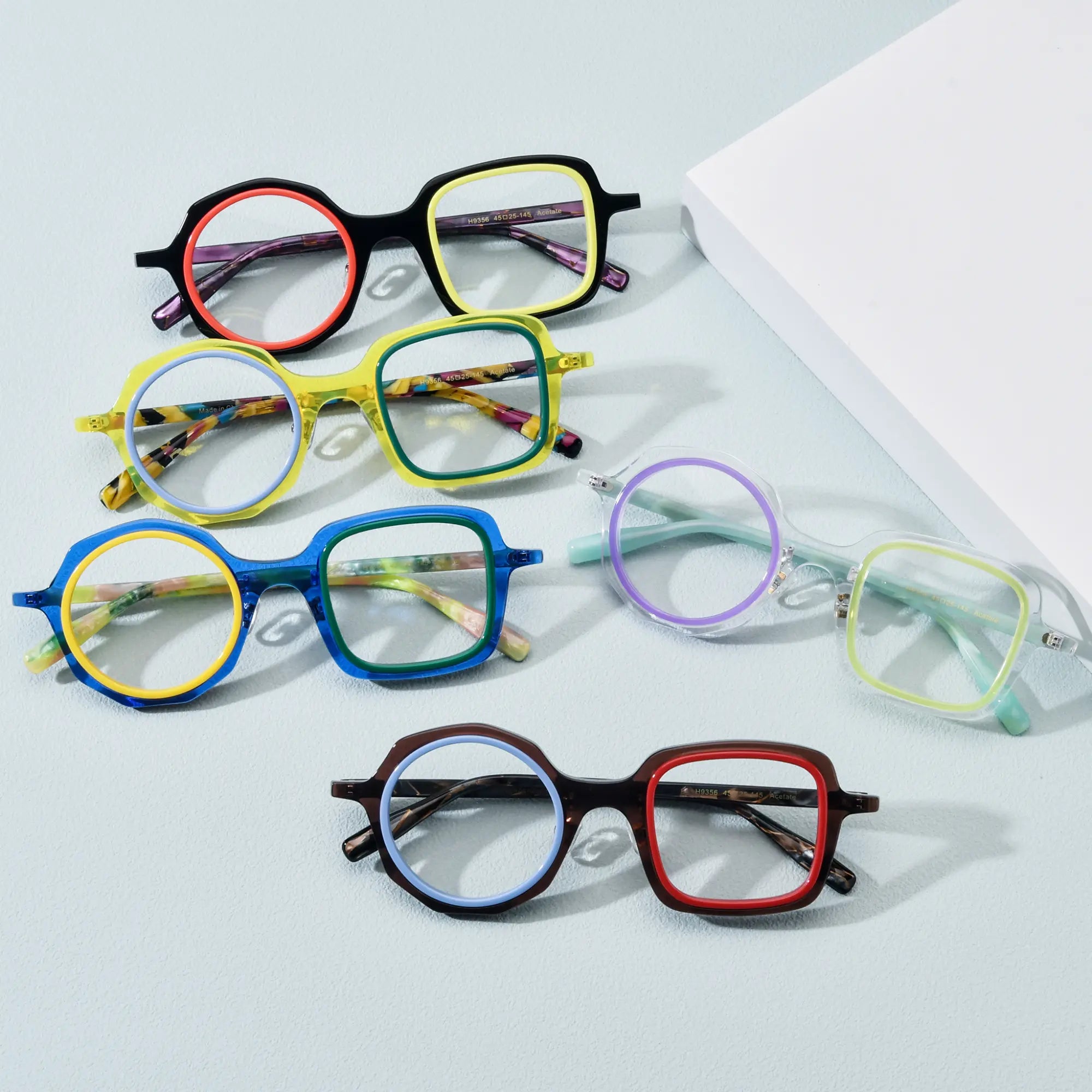 Flat lay of VoyueLens™ VL19356 eyeglasses in all five color variants: Black Amber, Honey Mint, Navy Lemon, Crystal Lilac, and Chestnut Blue. Displayed on a light blue background with acetate frames and dual-tone rims.
