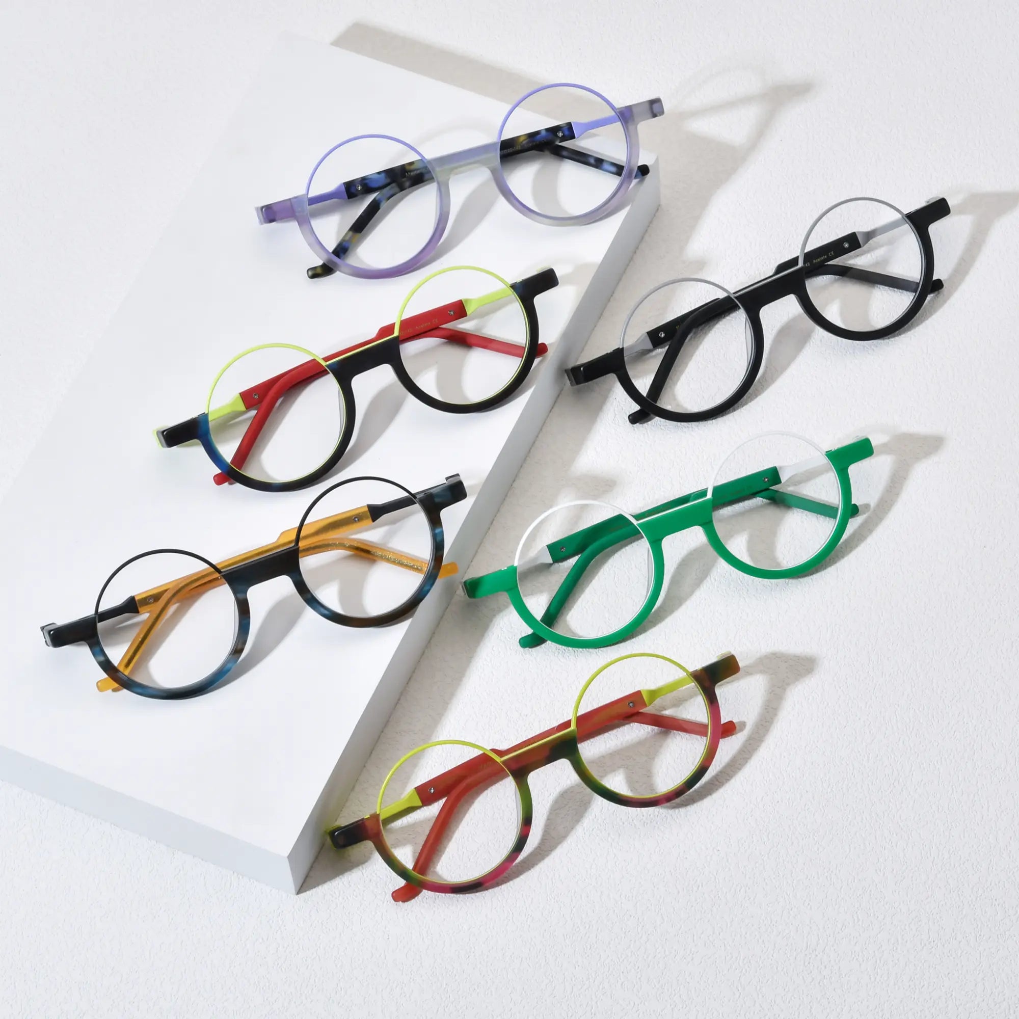 Top-down view of VoyueLens™ VL19366 round eyeglasses in six vibrant colorways, including Aurora Fade, Charcoal Silver, Citrus Fade, Ocean Tortoiseshell, Crimson Nebula, and Emerald Ice, arranged aesthetically on a white platform.