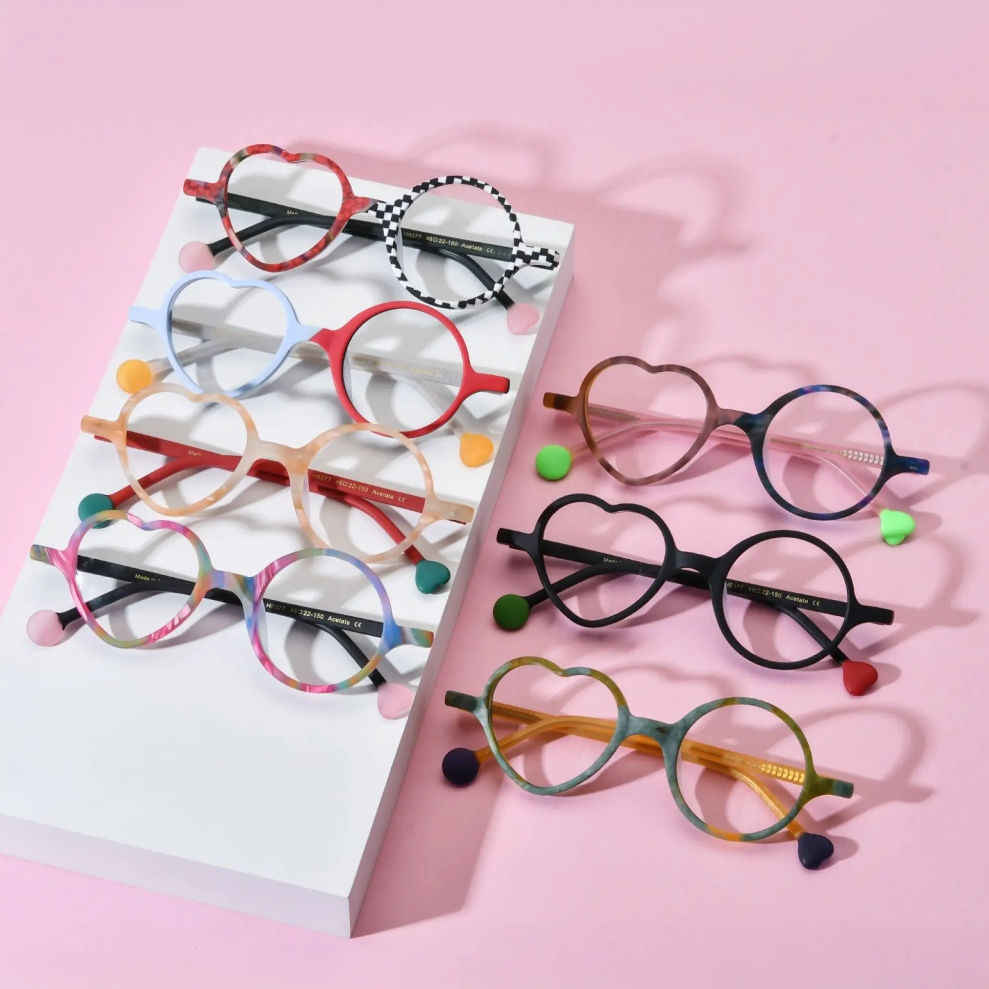 Group display of VoyueLens™ VL19377 Whimsy Pop Eyeglasses in all seven color variants, featuring asymmetric heart and round lenses on a pink background. Stylish matte acetate frames with vibrant tones including Checker Red Pop, Ice Blue Cherry, Honey Peach, Cocoa Marble, Citrus Jungle, Rainbow Twist, and Charcoal Dusk.